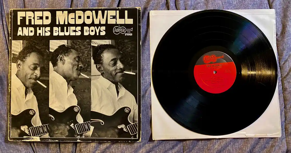 A vintage vinyl record and its album cover for 'Fred McDowell and His Blues Boys' are displayed side-by-side on a blue…