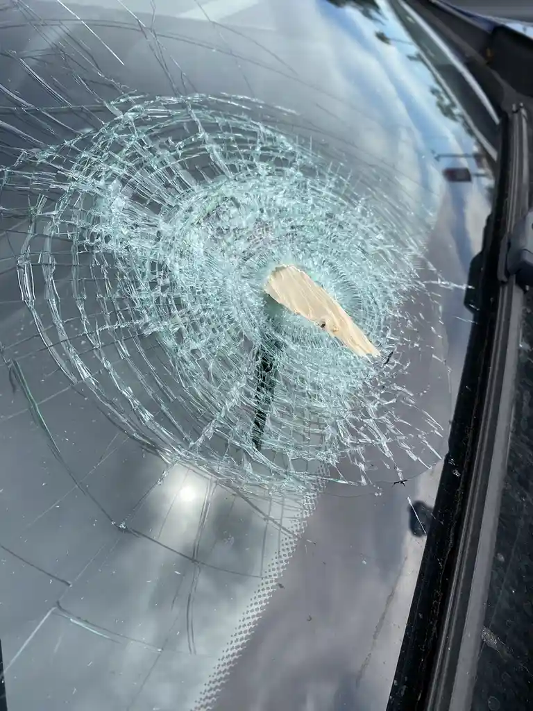 A close-up photo shows a car's windshield shattered in a circular pattern with a small wooden stick embedded in the center…