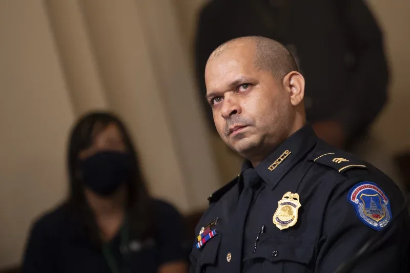 A man in a dark blue police uniform with visible badges and insignia stares intensely at the camera. Behind him, a woman…