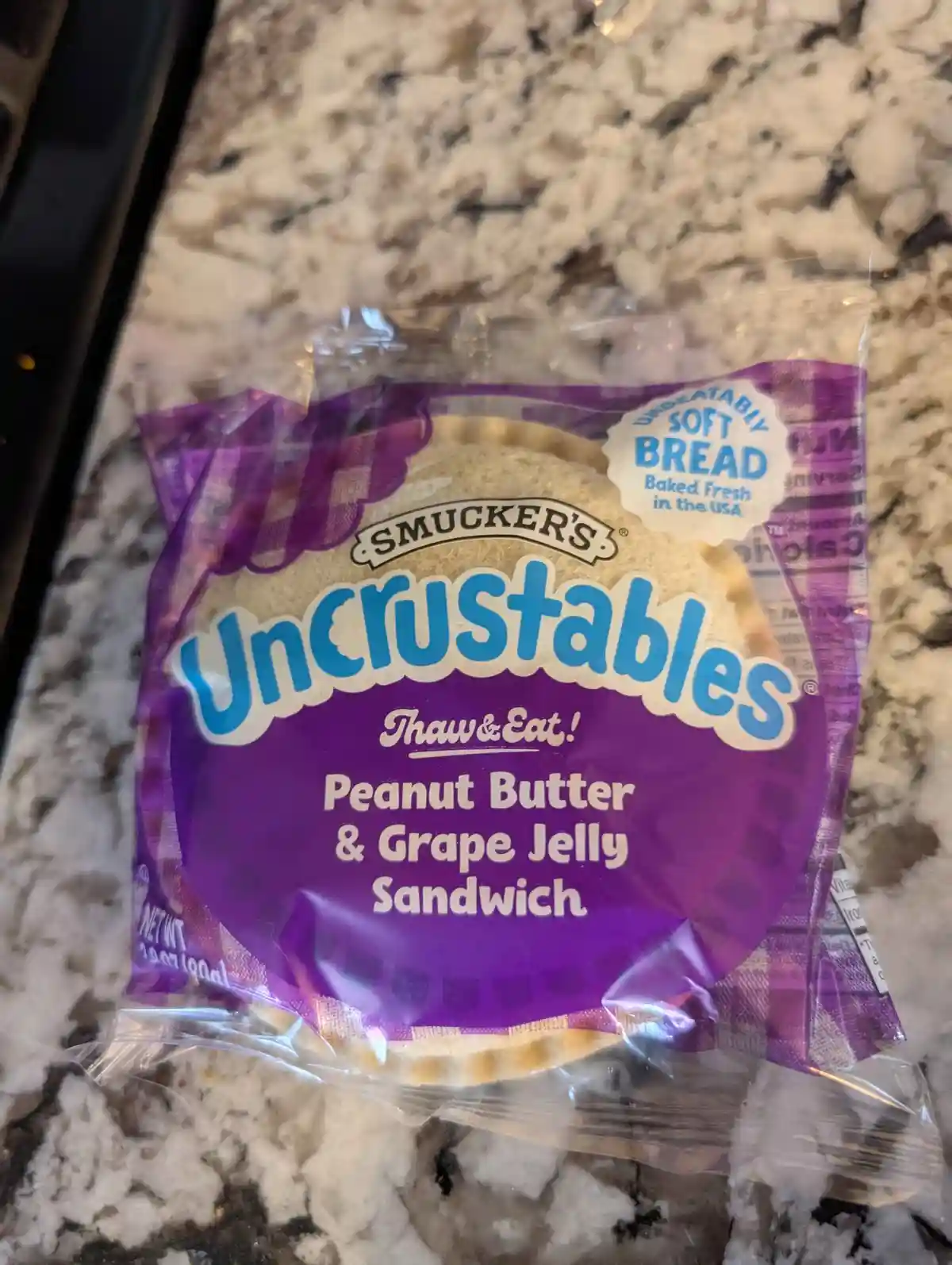 A package of Smucker's Uncrustables, specifically the peanut butter and grape jelly flavor, sits on a speckled countertop…