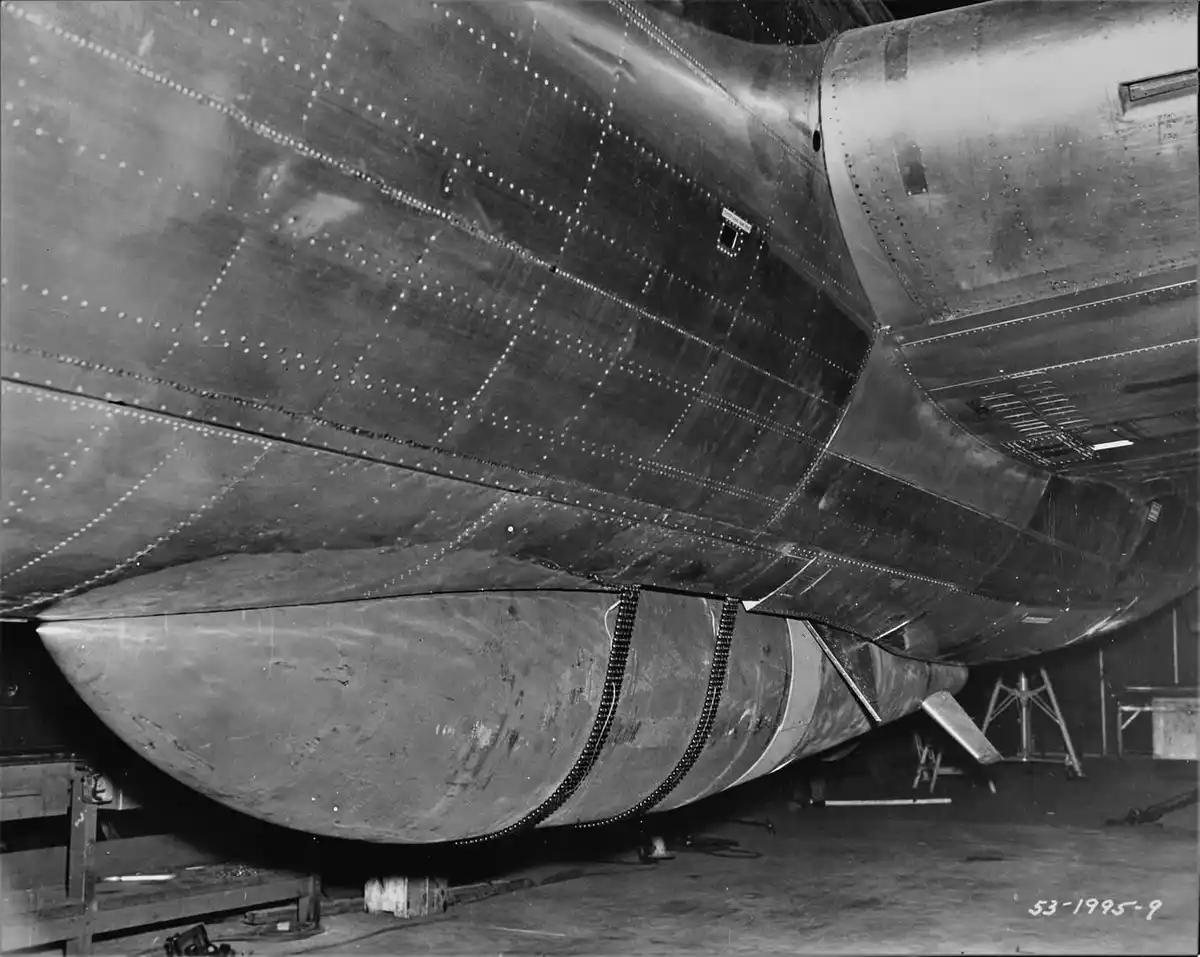 A black-and-white photograph shows the underside of a large, riveted metal aircraft fuselage in a hangar, with a prominent…