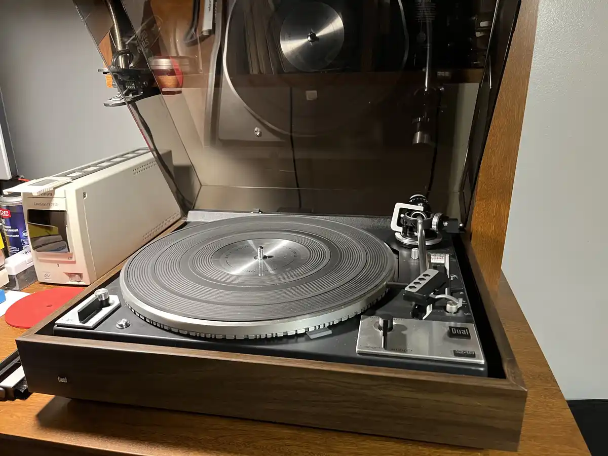 A vintage Dual 1249 turntable with its lid open, revealing an empty platter and tonearm, sits on a wooden desk next to a…