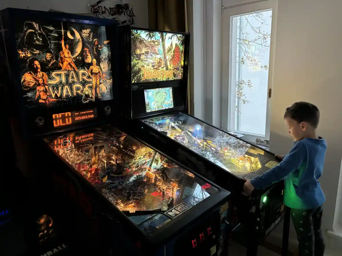 A young child in a blue shirt stands focused at a lit Jurassic Park pinball machine, with a Star Wars pinball machine beside…