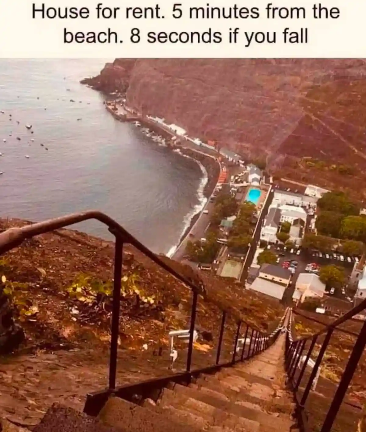 An aerial view shows a steep, narrow stone staircase with metal railings descending from a cliff edge toward a coastal town…