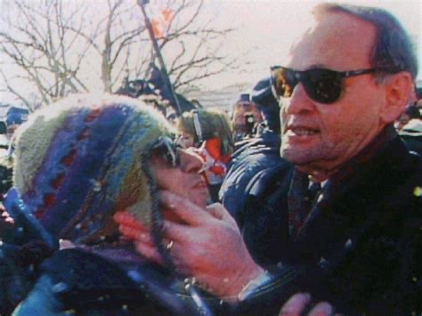 A man in sunglasses gently touches the cheek of a woman wearing a colorful knit hat and sunglasses, both smiling in a crowd…