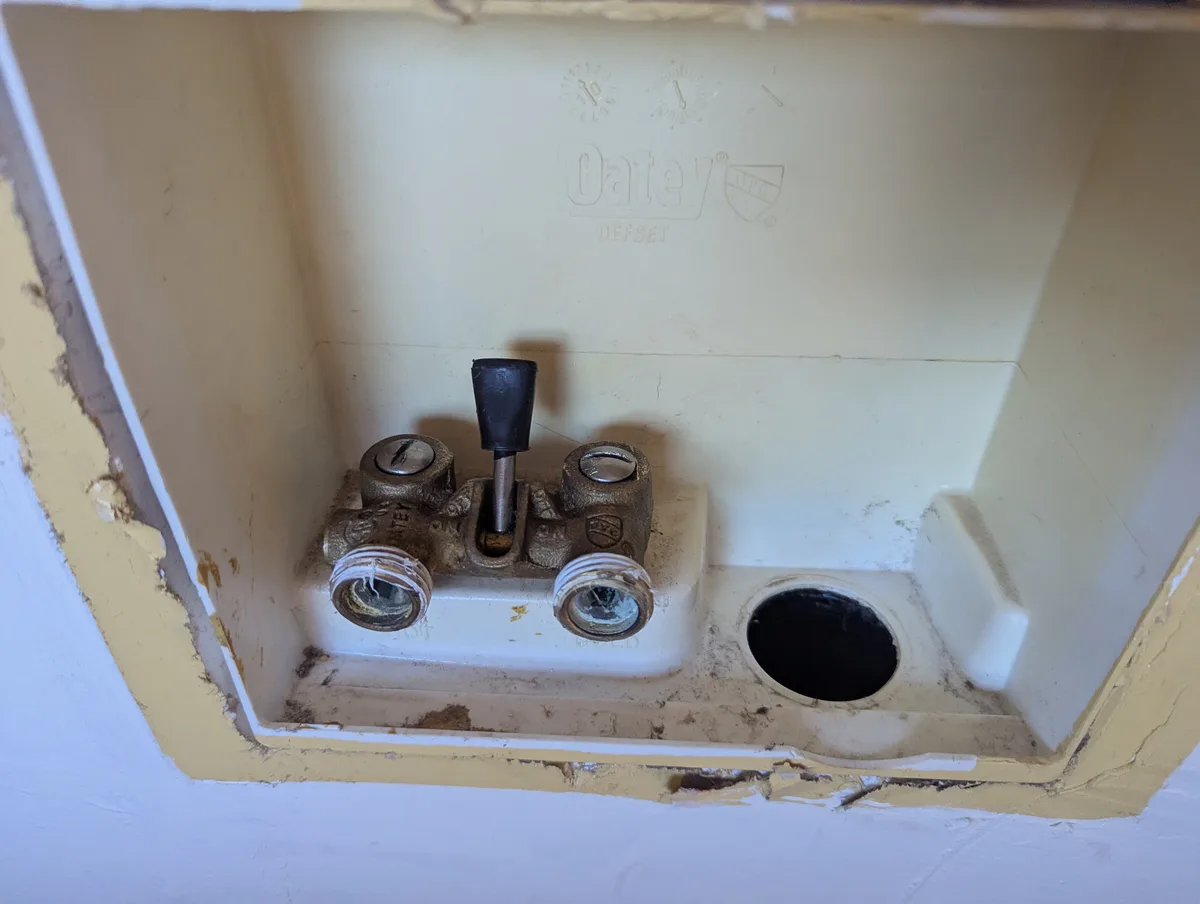 a close-up photo shows a rusty, old-fashioned Oatey brand plumbing valve inside a dirty, yellow-painted wall box. The valve…