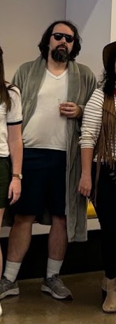 A man with long dark hair, a beard, and sunglasses stands in a group, wearing a white t-shirt, dark shorts, and a light…