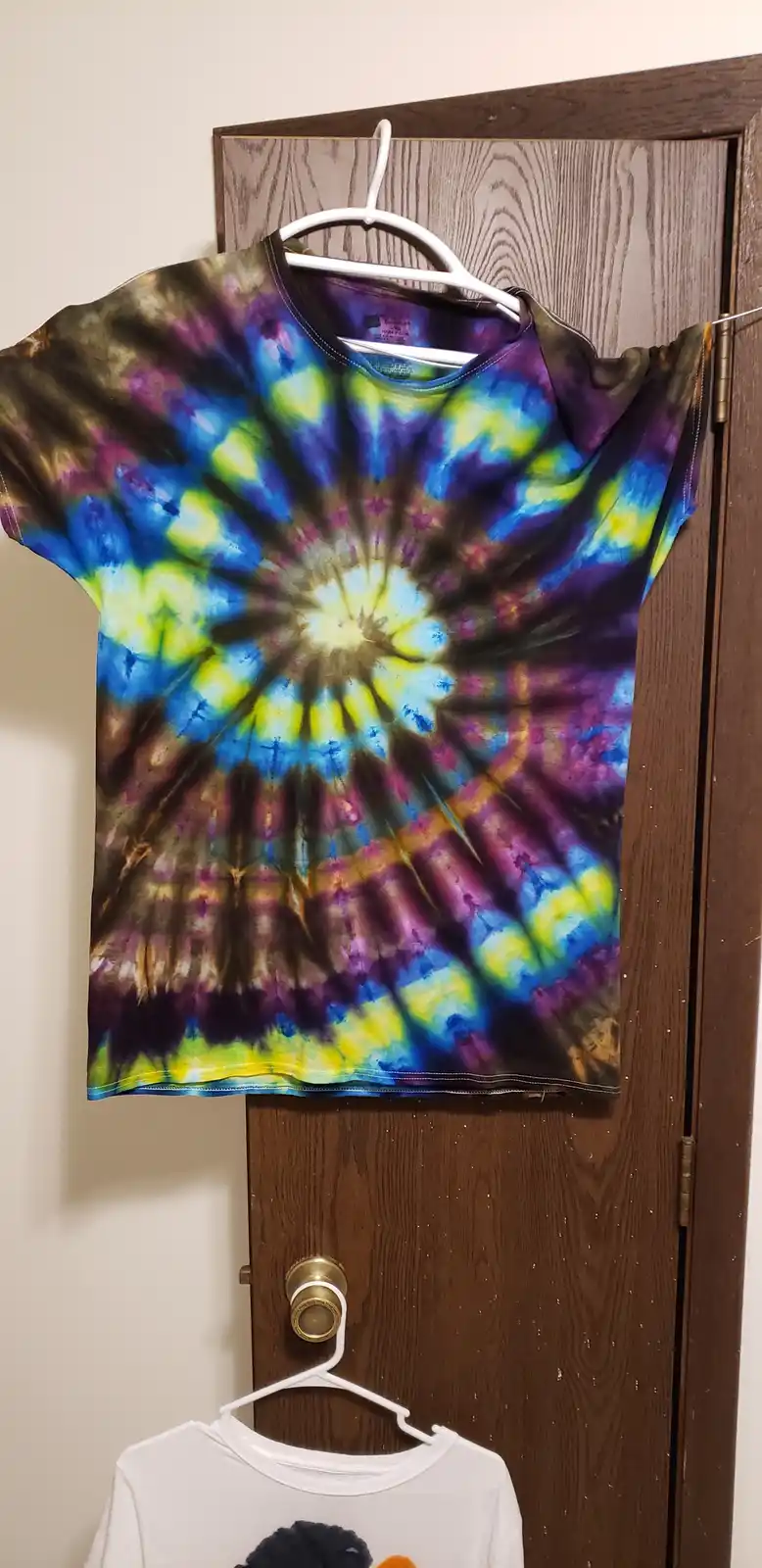 A vibrant, multi-colored spiral tie-dye t-shirt hangs on a white plastic hanger from a wooden door, with a plain white…