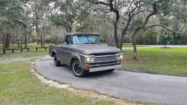 A vintage gray Ford F-100 pickup truck is parked on a paved road in a park with moss-draped trees. The truck's headlights…