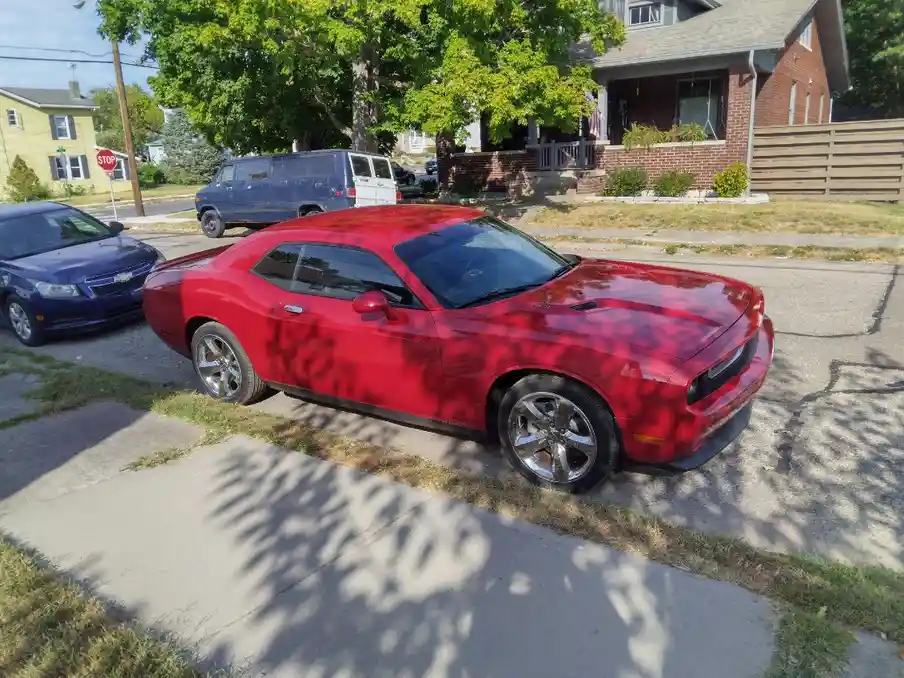 A red Dodge Challenger muscle car is parked on a residential street, its glossy paint reflecting tree shadows. The car is…