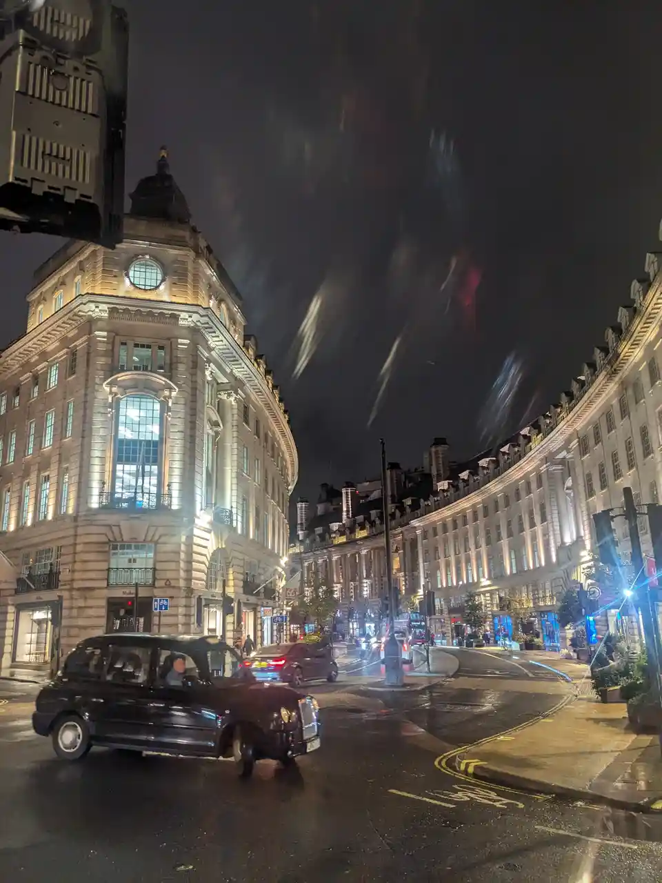 A nighttime photo of a wet London street corner with classic architecture, a black taxi in the foreground, and streaks of…