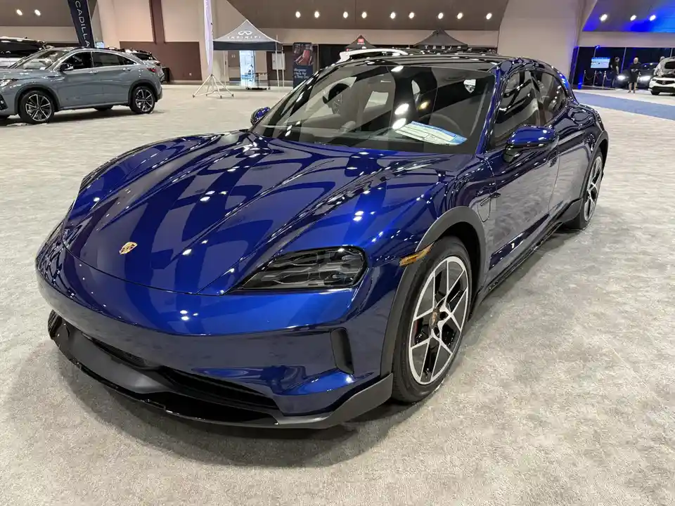 A glossy blue Porsche electric vehicle is displayed at an auto show, with other cars and brand tents visible in the…