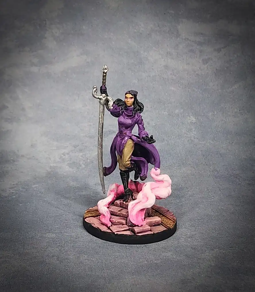 A painted miniature figurine of a female warrior in purple robes and black boots, standing on a circular base with pink…