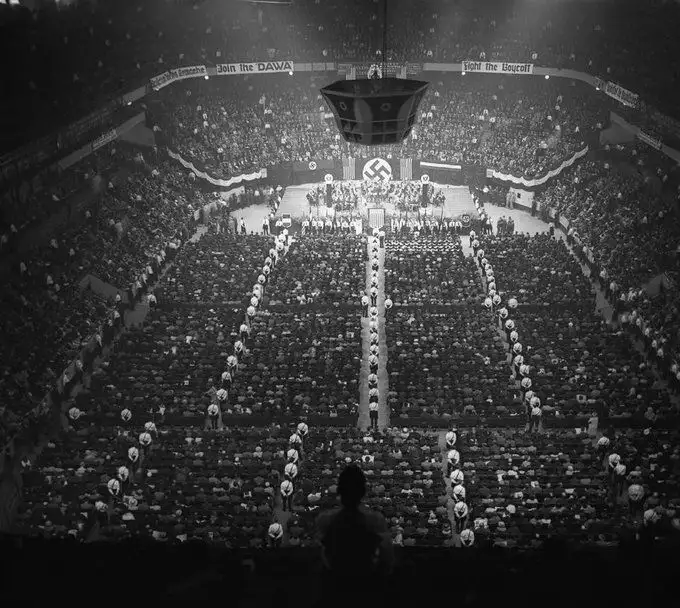 A black-and-white historical photograph shows a massive Nazi rally in a stadium, with rows of attendees and a stage…