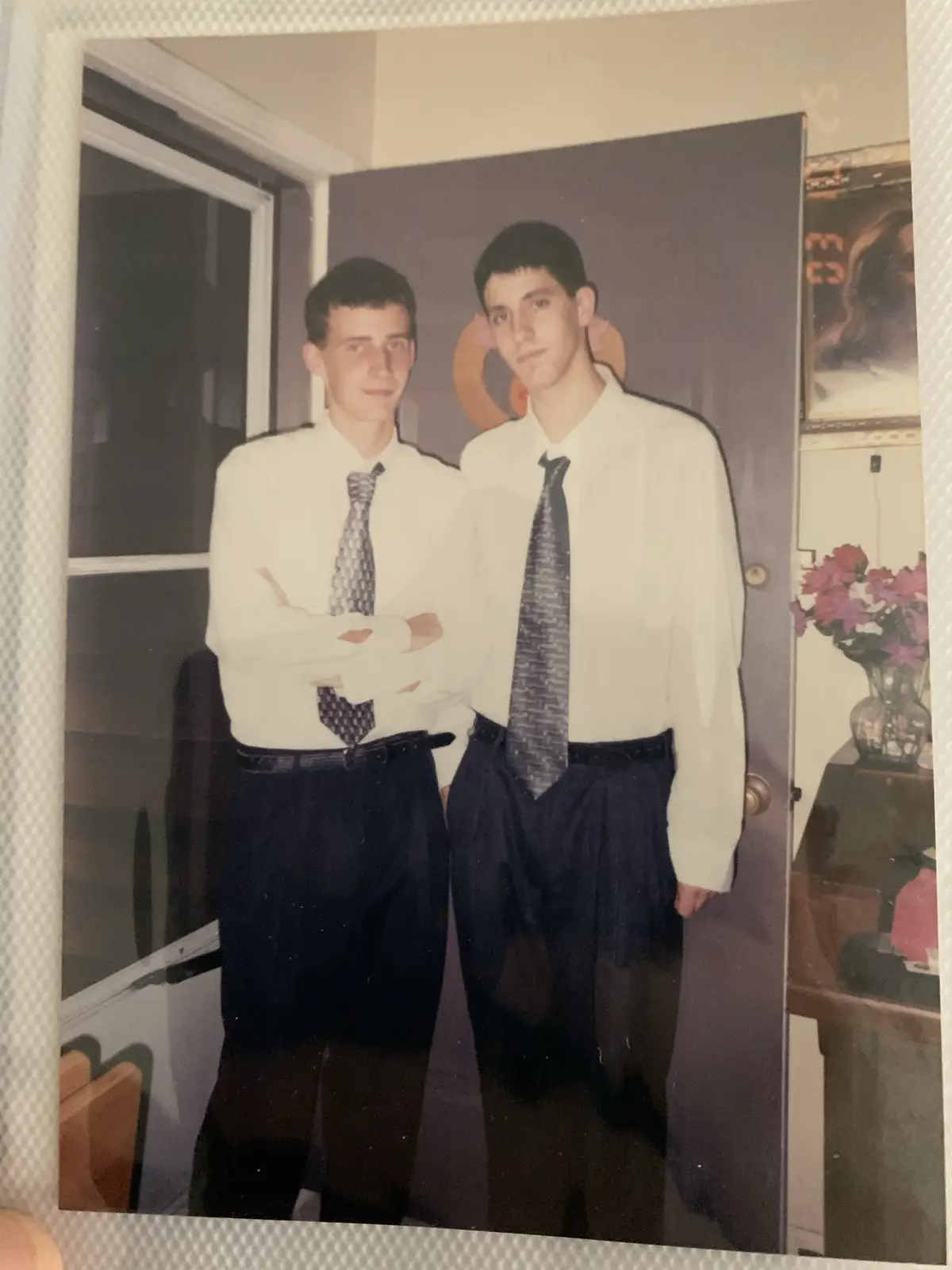 Two young men in matching white shirts and dark ties stand side by side indoors, one with arms crossed, the other with one…