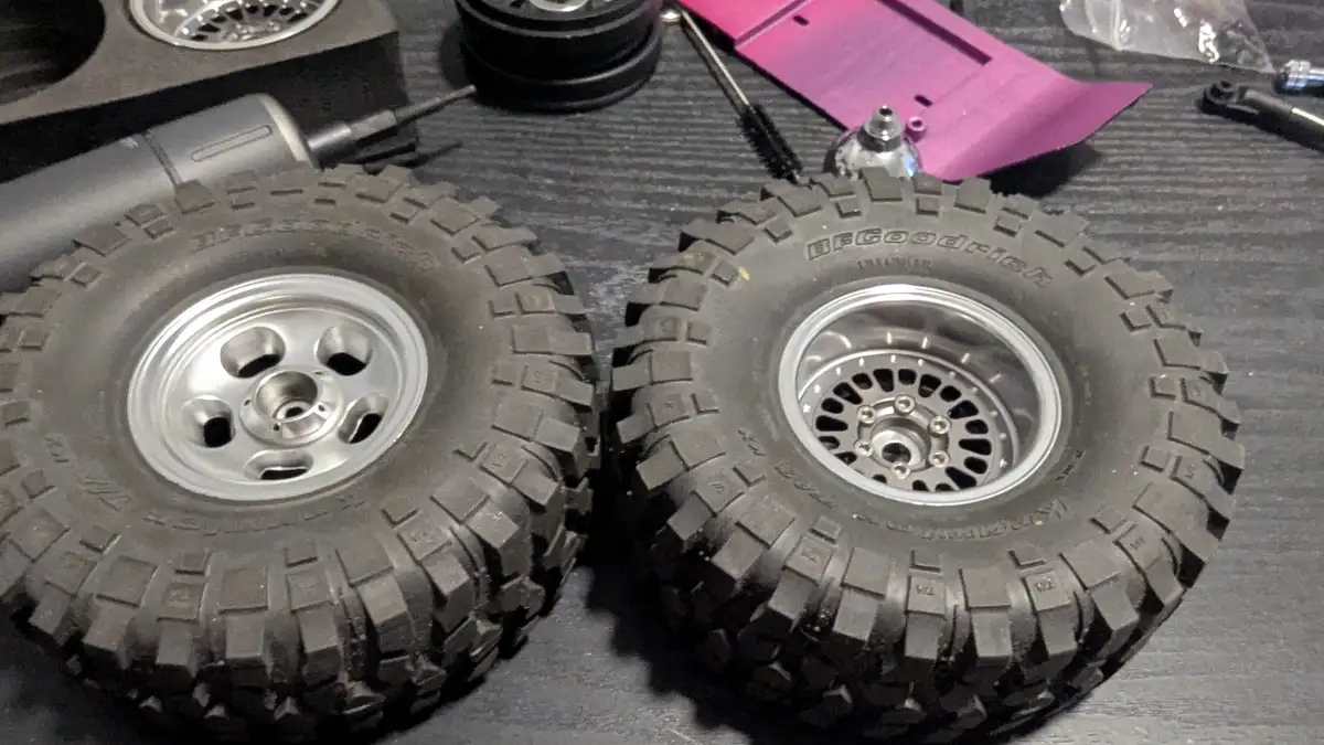 Two large, rugged RC car tires with silver rims sit side by side on a dark textured surface, surrounded by RC car parts…