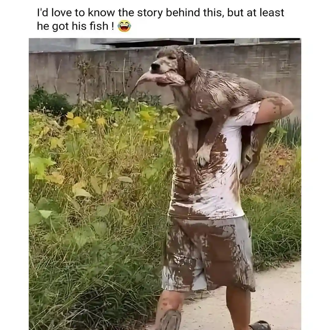 A person covered in mud carries a muddy dog on their shoulders; the dog has a fish stuck in its mouth. The person and dog…