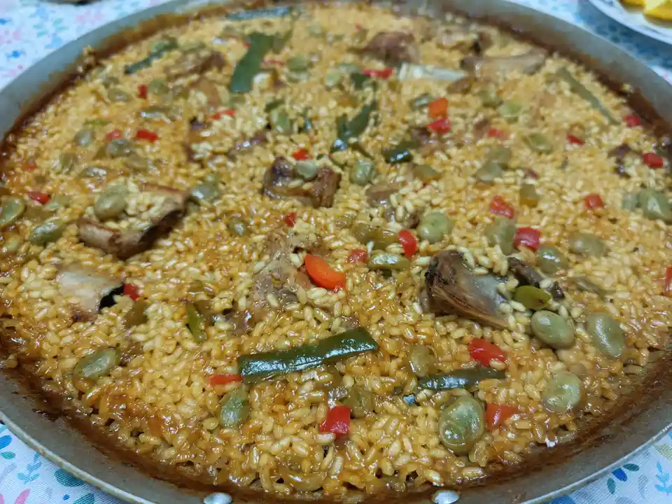 a close-up photo of a large, round metal pan filled with paella, featuring golden rice mixed with green beans, red bell…