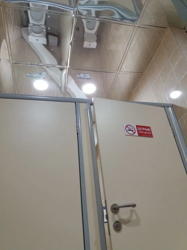 A low-angle photo of a public restroom stall door with a red sticker that reads 'ШТРАФ 1000 рублей' (Fine 1000 rubles) next…