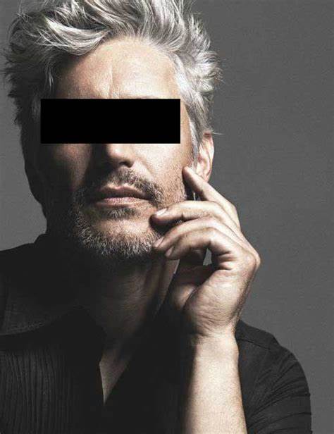 A man with gray hair and a beard, wearing a black shirt, has his eyes obscured by a black bar. His hand rests near his chin…