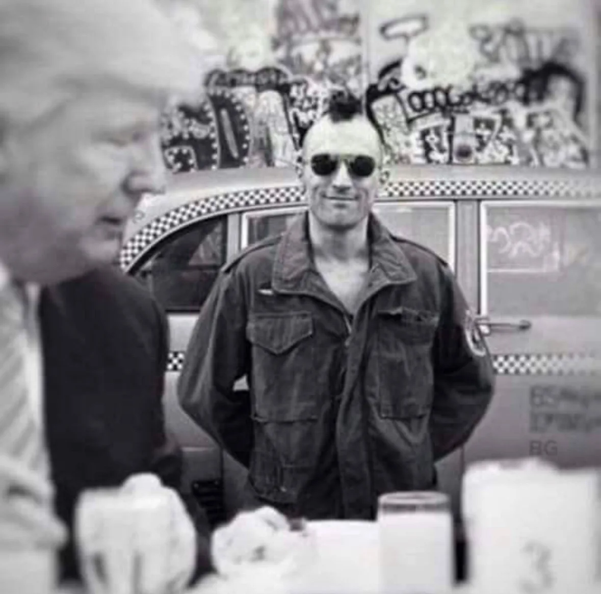 A black-and-white photo shows a man in a military-style jacket and sunglasses standing confidently in front of a taxi, while…